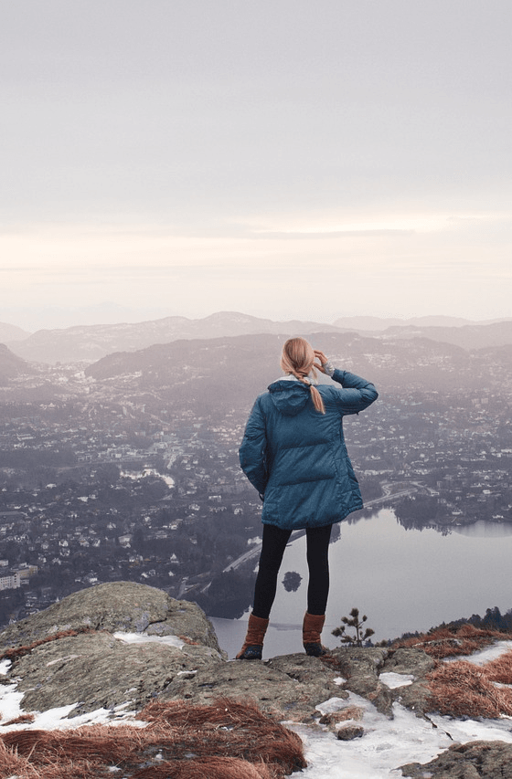 A woman stands on a mountain peak, her hand shades her eyes as she overlooks the valley below