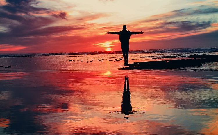 The silouette of a person with outstretched arms is seen standing along the water's edge, their reflection appearin below them as they raise their arms to the rising sun.