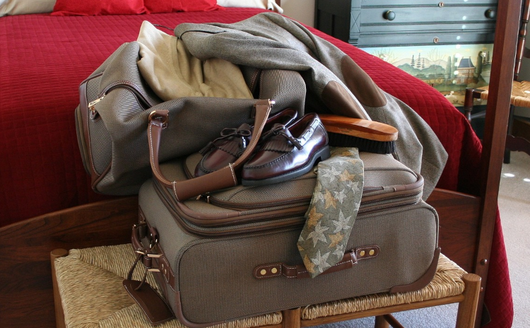 A heap of luggage sits on a bench at the end of a bed draped with a coat, shoes and other items, in preparation for a trip.