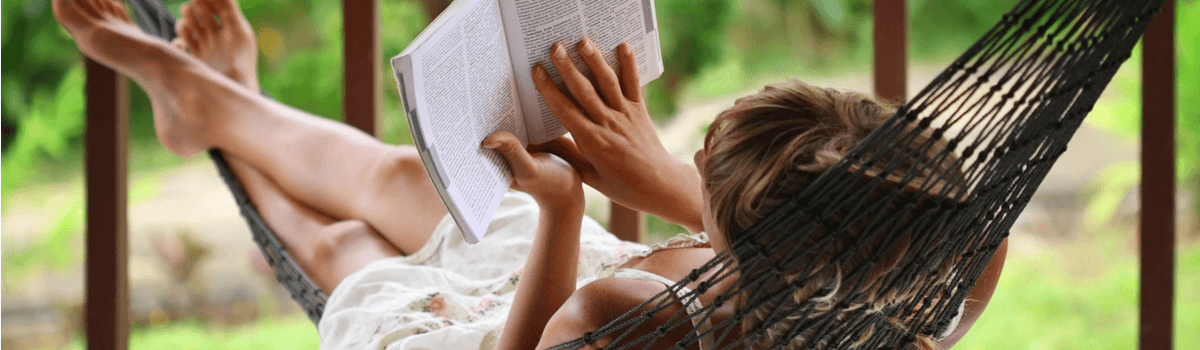 A woman swings in a hammock, while reading the open pages of a book. The view is from behind and slightly above her.