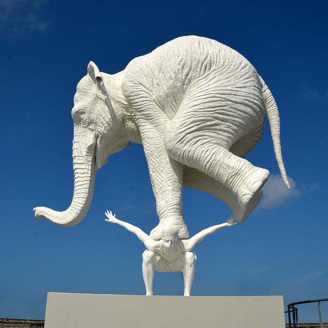 Full image - White statue of a man bent down, arms outstretched with a white elephant on his back with the elephant balancing on his front legs.